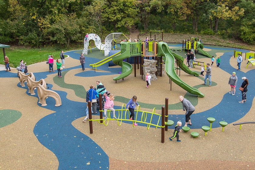 Large community playground with slides, climbers, and balance equipment installed over poured-in-place rubber safety surfacing, with families and children actively using the play area.